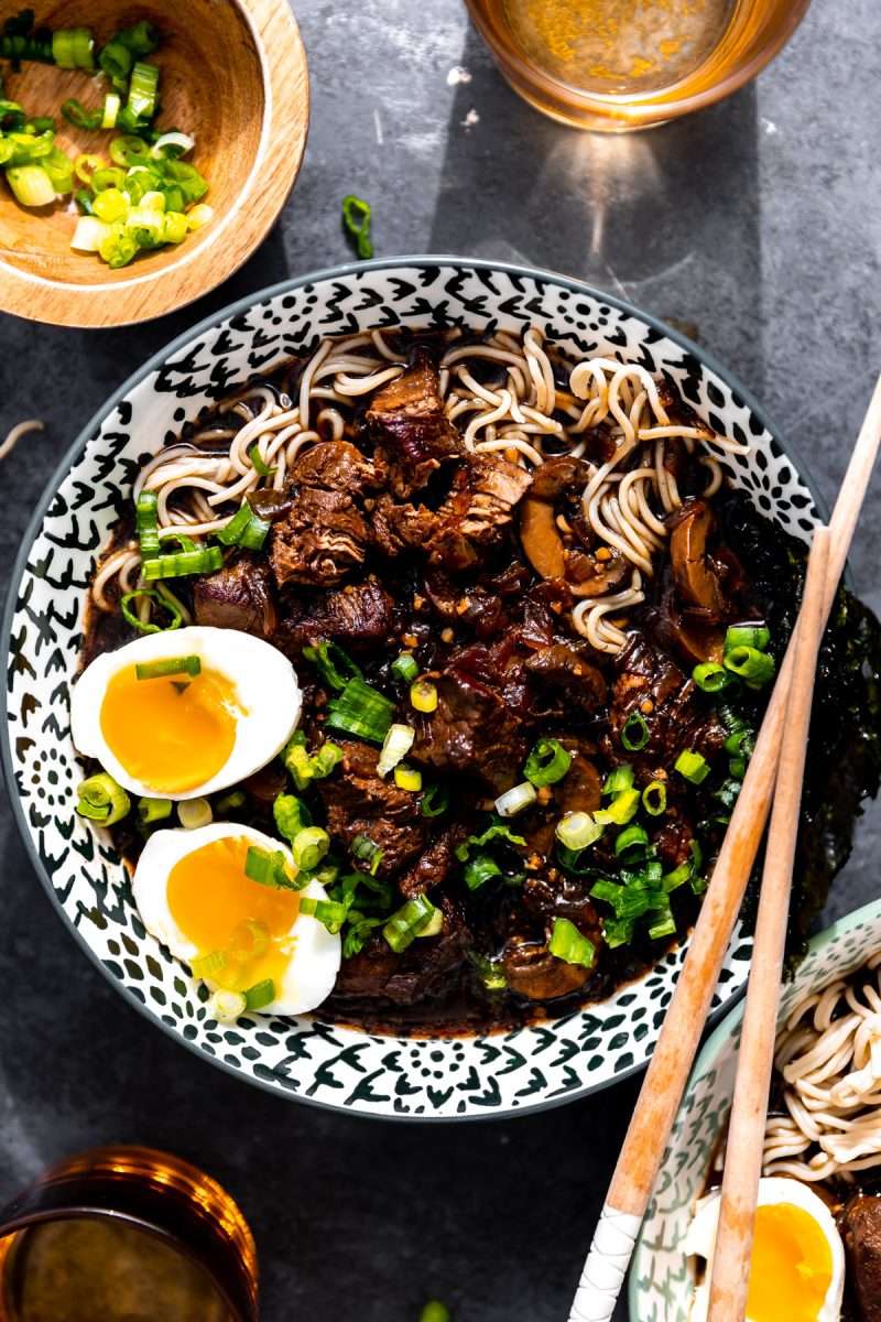 A bowl of beer braised beef ramen topped with soft boiled eggs and diced green onion.
