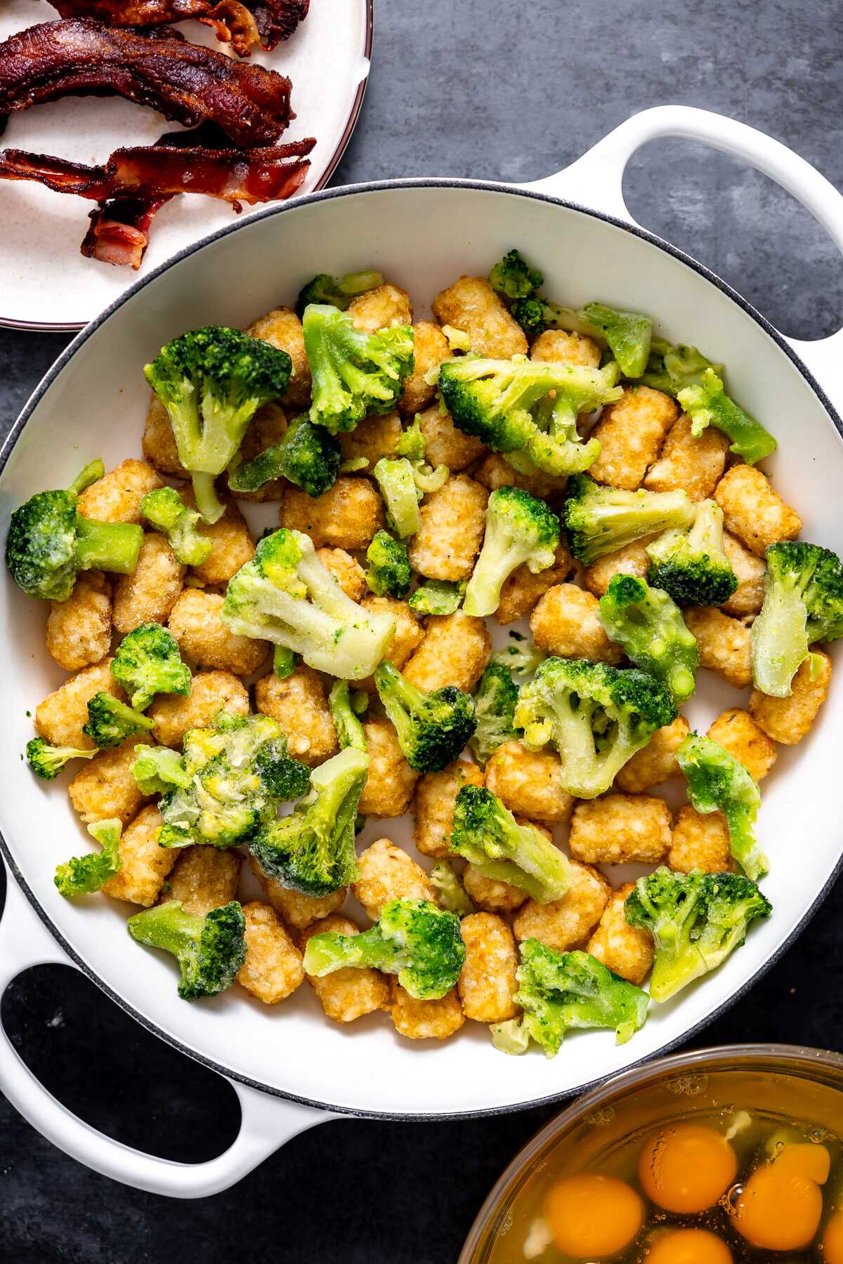 Frozen broccoli and tater tots in a baking dish with bacon and eggs waiting to be added on. 
