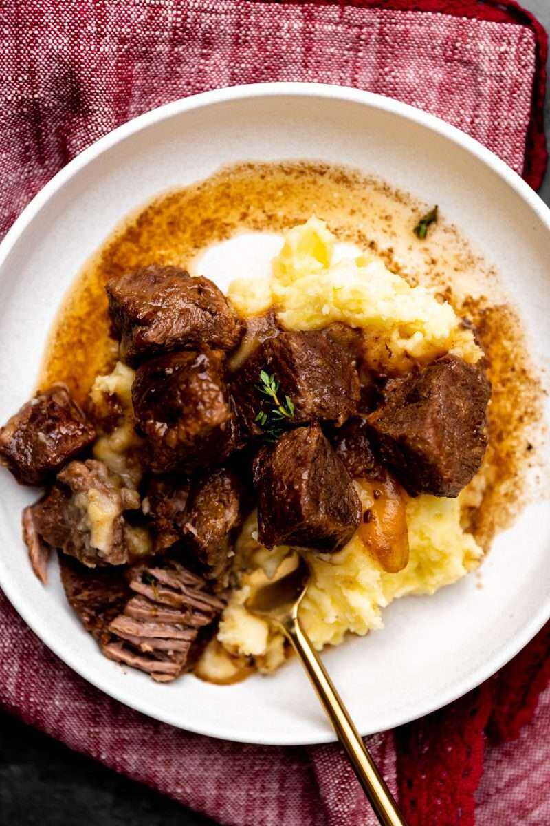 A plate of red wine braised beef tips on top of mashed potatoes.