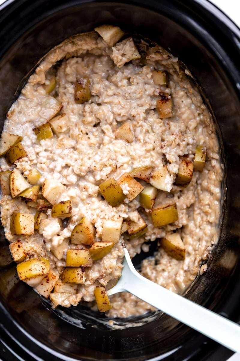 an overhead photo of a white spoon scooping up cooked oatmeal, apples from a crockpot. 