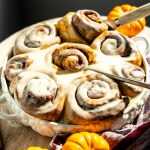 frosted pumpkin cinnamon rolls baked in a dish with a spatula pulling one out.